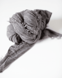 A close-up of the Grisal Love Cashmere Scarf in Dune, tied in a loose knot on a white surface, highlights its frayed edges and heirloom quality.