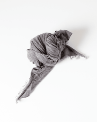 The Grisal Love Cashmere Scarf in Dune, in soft gray cashmere, lies crumpled on a white surface, loosely knotted at the center with frayed, trailing ends.