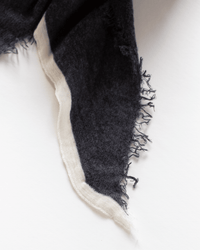 A close-up of the Grisal Rosa Cashmere Scarf in Charcoal X Milk shows its frayed, dark edge with a white border on a white background, highlighting the unique texture and elegance of this accessory.