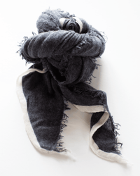 The Grisal Rosa Cashmere Scarf in Charcoal X Milk, featuring white fringed edges, is loosely knotted and elegantly arranged on a white background.