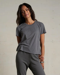 A woman with long dark hair smiles and poses in the perfectwhitetee Harley Cotton S/S Boxy Crew Tee in Charcoal and matching gray pants, standing against a plain, light-colored wall.