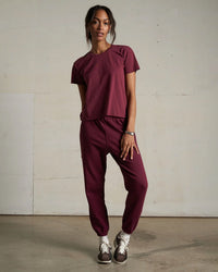 A woman stands indoors by a plain wall, wearing perfectwhitetee’s Johnny French Terry Easy Sweatpant in Dark Burgundy with brown sneakers. She rests one hand on her thigh and gazes slightly to the side.