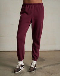 A person stands on a concrete floor against a plain light wall, wearing perfectwhitetee’s Johnny French Terry Easy Sweatpant in Dark Burgundy, white socks, and black-and-white Nike sneakers. Their torso and head are not visible.