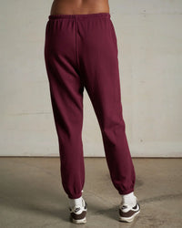 A person stands indoors on a concrete floor, wearing perfectwhitetee’s Johnny French Terry Easy Sweatpant in Dark Burgundy, paired with white socks and Nike sneakers. The waist-down photo is taken from behind.