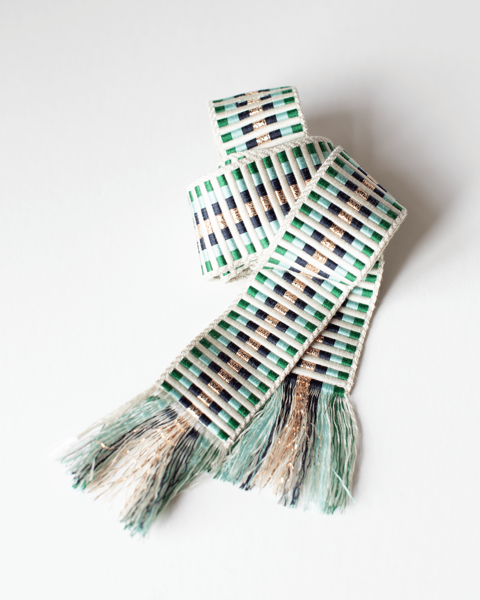 The Alona Woven Belt in Lurex & Green by Hartford features green, black, and beige stripes with fringed ends, displayed on a white surface to highlight its subtle geometric pattern.