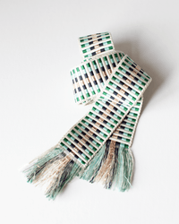 The Alona Woven Belt in Lurex & Green by Hartford features green, black, and beige stripes with fringed ends, displayed on a white surface to highlight its subtle geometric pattern.