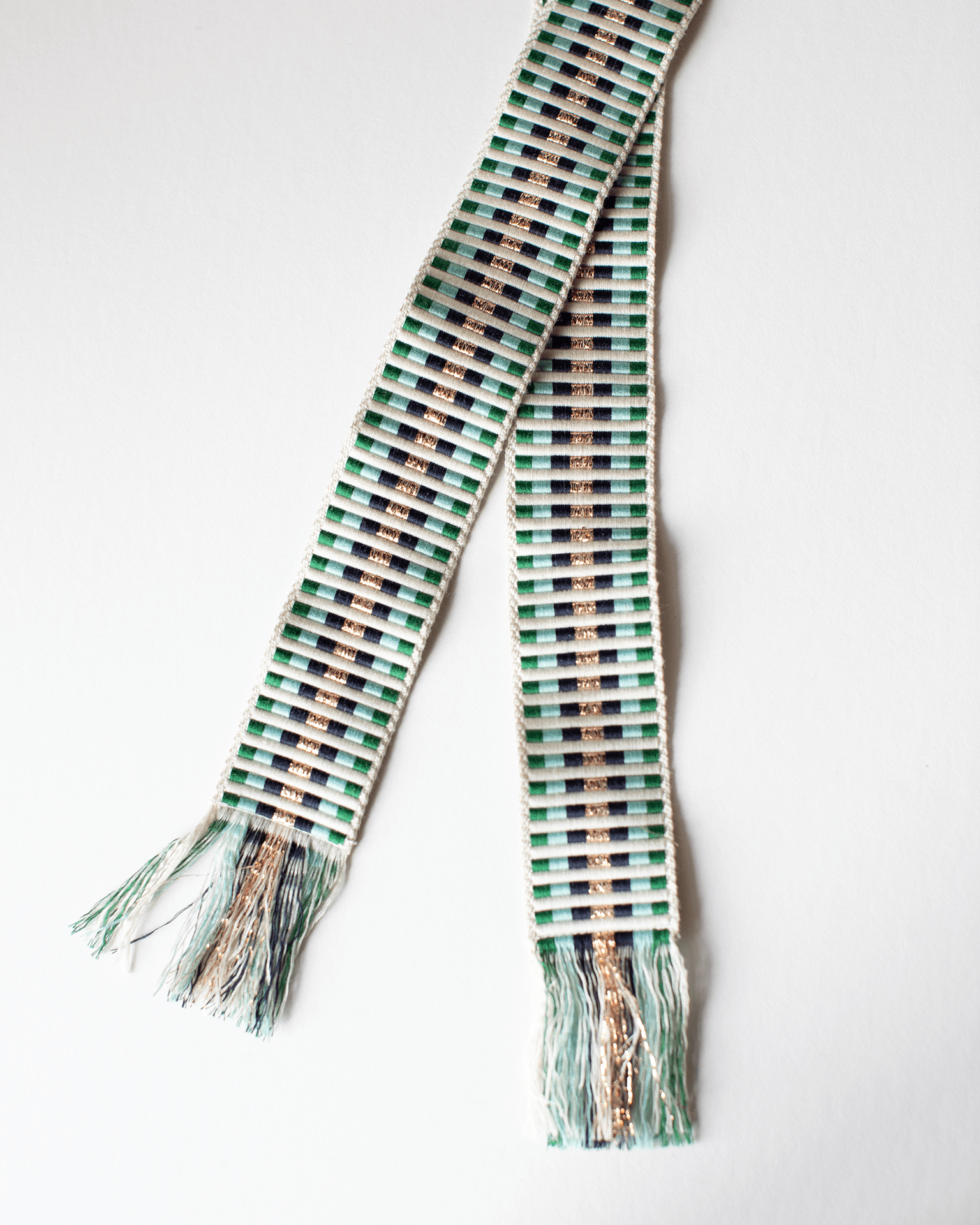 The Hartford Alona Woven Belt in Lurex & Green features green, white, black, and metallic gold stripes with a subtle geometric pattern and fringed ends, displayed on a white background.