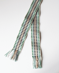 The Hartford Alona Woven Belt in Lurex & Green features green, white, black, and metallic gold stripes with a subtle geometric pattern and fringed ends, displayed on a white background.