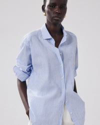 A person with short hair and gold hoop earrings stands against a plain background wearing the Hartford Charlot Shirt in White & Blue, a loose-fitting cotton seersucker top with stripes, rolled-up sleeves, and side pockets.