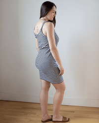 A woman with long brown hair stands indoors against a white wall and wooden floor, wearing Hartford’s Tella Striped Tank Dress in Ecru & Navy and brown sandals. She is turned to the side, showing a tattoo on her upper back.