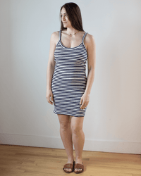 A woman with long brown hair stands indoors on a wooden floor, wearing the Hartford Tella Striped Tank Dress in Ecru & Navy and brown sandals. She looks slightly to the side with a neutral expression.