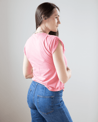 A woman with long brown hair, wearing the Hartford Tomari V Neck Sleeveless Top in Hibiscus and blue jeans, stands turned slightly away from the camera against a plain light background.