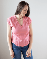 A woman with long brown hair wears the Hartford Tomari V Neck Sleeveless Top in Hibiscus and blue jeans, standing against a plain light background while looking down with a slight smile.