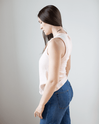 A woman with long dark hair stands sideways, looking down. She wears a sleeveless Tulette Henley Tank in Blush by Hartford and blue jeans, set against a plain light background.