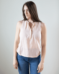 A woman with long brown hair wears the Hartford Tulette Henley Tank in Blush, a sleeveless light pink top, paired with blue jeans. She stands against a plain background, gazing to the side with a neutral expression.