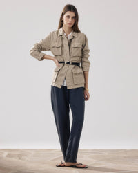A woman stands on a stone floor wearing the Hartford Vea Jacket in Straw—a belted, military-inspired style—paired with a light shirt, dark loose trousers, and black sandals. She has long hair and minimal accessories against a plain background.