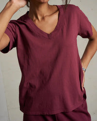 Wearing the perfectwhitetee Hendrix Cotton S/S Boxy V Neck Tee in Dark Burgundy and matching pants, a person stands with one hand on their hip and the other arm bent, face partially out of view.