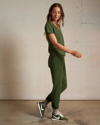 A woman with long brown hair wears an olive green V-neck top and the Johnny French Terry Easy Sweatpant in Fern Green by perfectwhitetee. She completes her look with gray socks, green Nike sneakers, and walks indoors against a neutral wall.
