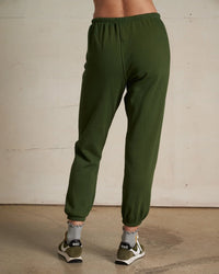 A person stands indoors facing away, wearing the Johnny French Terry Easy Sweatpant in Fern Green by perfectwhitetee, paired with white Nike sneakers and gray socks. The setting features a light wall and concrete floor.