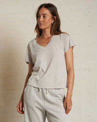 A woman wears the perfectwhitetee Hendrix Cotton S/S Boxy V Neck Tee in Silver, paired with matching drawstring pants, as she stands against a plain light-colored wall with a relaxed expression.