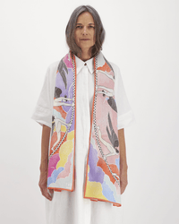 A woman with long gray hair wears a loose white button-up dress and an Inoui Editions Scarf 100 Yoga in Multicolor, featuring vibrant patterns and abstract figures, standing against a plain white background.