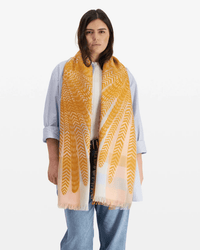 A person in a white shirt and blue jeans stands by a white background, draped in an Inoui Editions Scarf / Etole 70 3D Hedwige in Pastel—a large yellow and white cotton wool blend scarf with fringed edges.