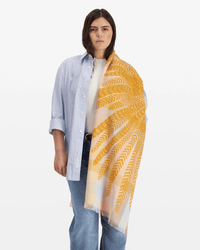 A person with straight brown hair wears a light blue striped shirt over a white top and blue jeans, styled with the Inoui Editions Scarf / Etole 70 3D Hedwige in Pastel draped over one shoulder.