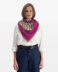 A woman with straight gray hair wearing a white button-up, blue jeans, and a black belt stands facing forward. She has the Inoui Editions Square/Carre 100 Chatou Winter in Purple scarf tied around her neck.