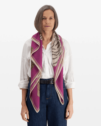 A woman with straight gray hair faces forward, wearing a white button-up shirt, dark blue high-waisted pants, and an Inoui Editions Square / Carre 100 Chatou Winter in Purple draped around her neck.