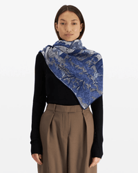 A woman faces forward in a black long-sleeve top, high-waisted brown trousers, and the Inoui Editions Square / Carre 100 Reve in Blue scarf draped over her shoulders. The background is plain white.