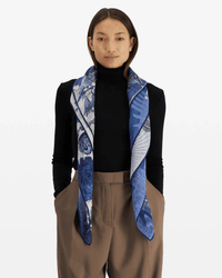 A person with straight brown hair wears a black turtleneck, brown trousers, and the Inoui Editions Square / Carre 100 Reve in Blue scarf, draped around their neck, standing against a plain white background.