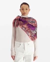A person wears a light beige turtleneck and white pants, with the Inoui Editions Square / Carre 100 Reve in Fuchsia—an oversized bandana—in pink and purple draped over one shoulder, standing against a plain white background.