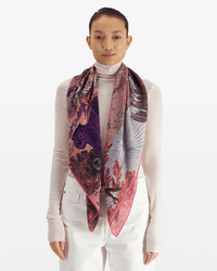 A person in a sheer white turtleneck and pants is draped with the Square / Carre 100 Reve in Fuchsia by Inoui Editions—a luxurious wool-silk-cashmere oversized bandana featuring pink, purple, and black designs with pink edging.