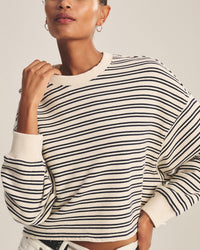 A person wearing the Isabelle Long Sleeve Crew Neck Top in Natural/Navy by Velvet by Graham & Spencer—a cream, cropped, boxy crewneck with black stripes—paired with hoop earrings and white pants. Their head is partly out of frame; hand touches collar.