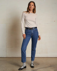 A woman stands indoors against a plain light wall, smiling with one hand in her pocket. She wears the perfectwhitetee Jamie Recycled Cotton L/S in Silver, blue high-waisted jeans, a black belt, black Mary Jane shoes, and light socks.