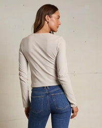 A woman with long brown hair faces away, wearing the perfectwhitetee Jamie Recycled Cotton L/S in Silver and blue high-waisted jeans made from recycled cotton, set against a plain off-white background.