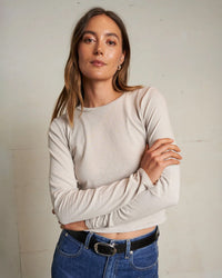 A woman with light brown hair wears the perfectwhitetee Jamie Recycled Cotton L/S in Silver, paired with blue jeans. She stands against a light wall, arms crossed, looking confidently at the camera.