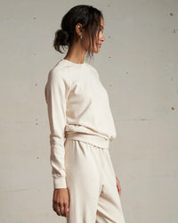 A woman in profile stands against a light wall, wearing the perfectwhitetee Alexi French Terry Sweatshirt in Sugar with matching sweatpants. Her dark hair is tied back in a loose bun.