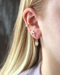 A close-up of a person with blonde hair in a lavender top, wearing Kris Nations' Safety Pin Enamel Hoop Earrings in White & 18K Gold Vermeil, along with other gold earrings.