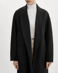 A person wearing the Lamarque Thara Jacket in Black, a long wool coat with oversized sleeves, layered over a white ribbed turtleneck and beige pants, shown from shoulders to thighs with hands by their sides.