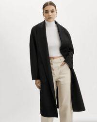 A woman with straight, slicked-back hair and gold hoop earrings wears the Lamarque Thara Jacket in Black over a white ribbed turtleneck crop top and high-waisted beige pants, standing against a plain background.
