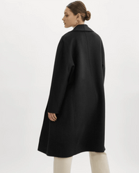 A woman with brown hair in a low bun wears the Lamarque Thara Jacket in Black, featuring oversized sleeves, over light-colored pants. She stands against a plain white background, facing away and looking over her shoulder.