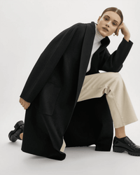 A woman kneels confidently, wearing the Lamarque Thara Jacket in Black over a white turtleneck and beige pants. Her hair is slicked back, and she completes the look with black chunky loafers.