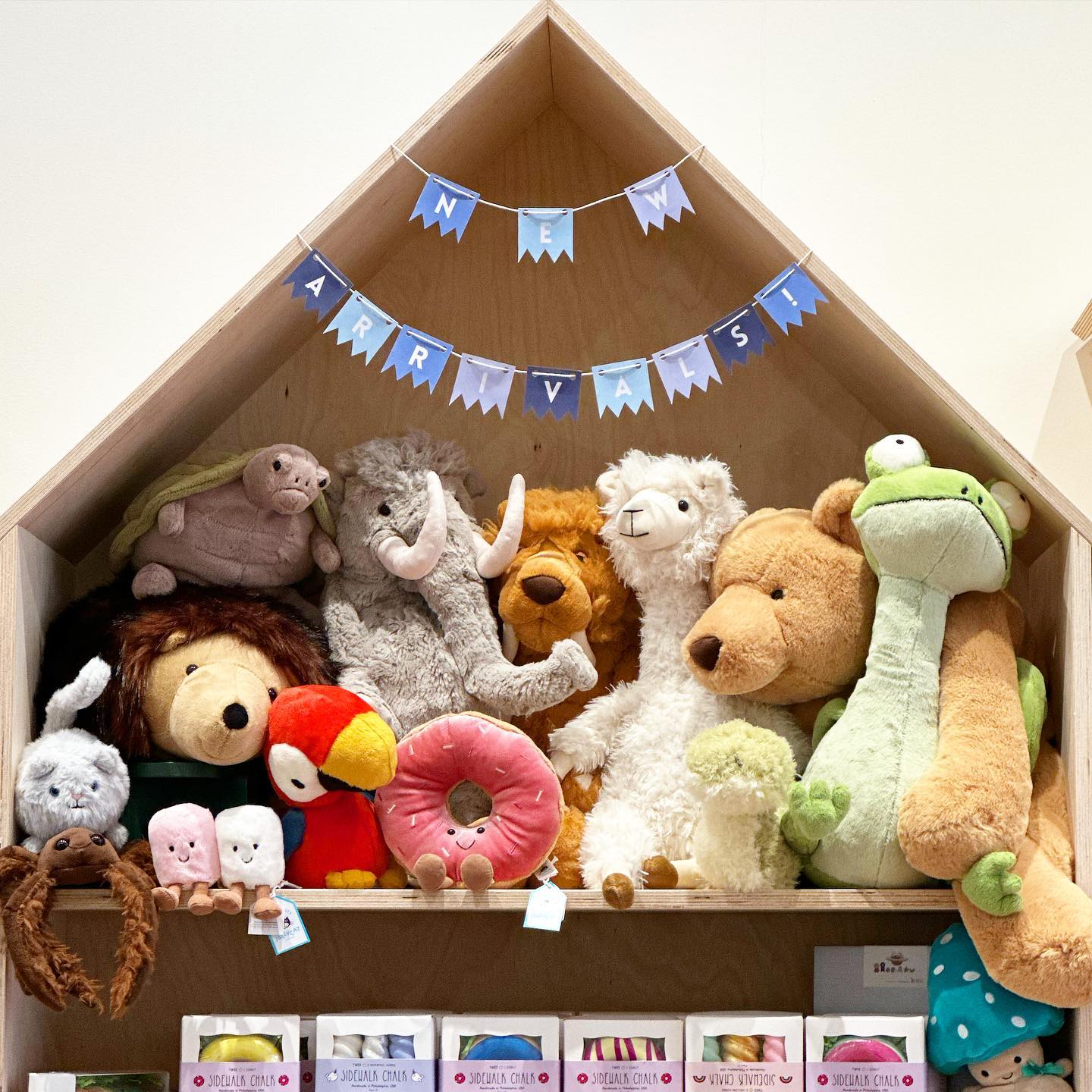 A display of various stuffed animals arranged on shelves inside a wooden house-shaped structure with a banner above reading "new arrivals".