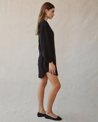 A woman stands in profile against a neutral background wearing the Bella Dahl Long Sleeve Belted Mini Dress in Black with black flats. Her hair is down and she faces left.
