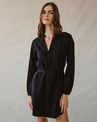 A woman with long brown hair wears a Bella Dahl Long Sleeve Belted Mini Dress in Black, standing against a light neutral background and looking confidently at the camera.