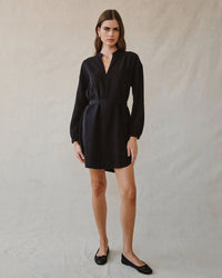 A woman with straight brown hair and a neutral expression stands against a plain beige background, wearing the Bella Dahl Long Sleeve Belted Mini Dress in Black and black flats, exuding effortless elegance.