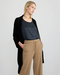 A woman in a black open cardigan, dark satin top, and beige wide-leg pants with front pockets stands with one hand in her pocket, gazing to the side—her look finished with the Travel Wrap in Black by Margaret O'Leary.