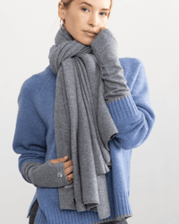 A woman wearing a light blue sweater, grey fingerless gloves, and a Margaret O'Leary Travel Wrap in Derby stands against a plain background, looking at the camera with a relaxed expression.