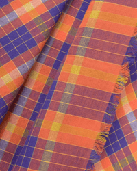 Close-up of the Moismont Plaid Bandana in Poppy Red, featuring colorful orange, yellow, blue, and red squares with intersecting lines. The oversized cotton scarf is folded to show a frayed edge on the right.